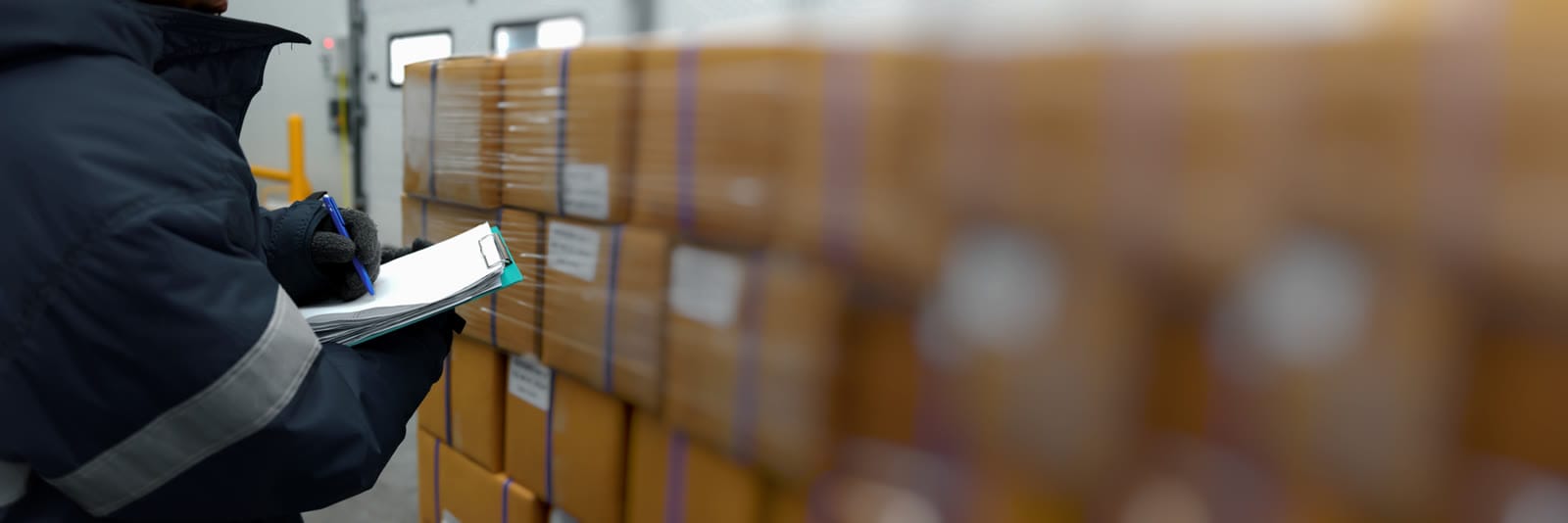 Warehouse hand checking an order against a clipboard beside stacked cartons